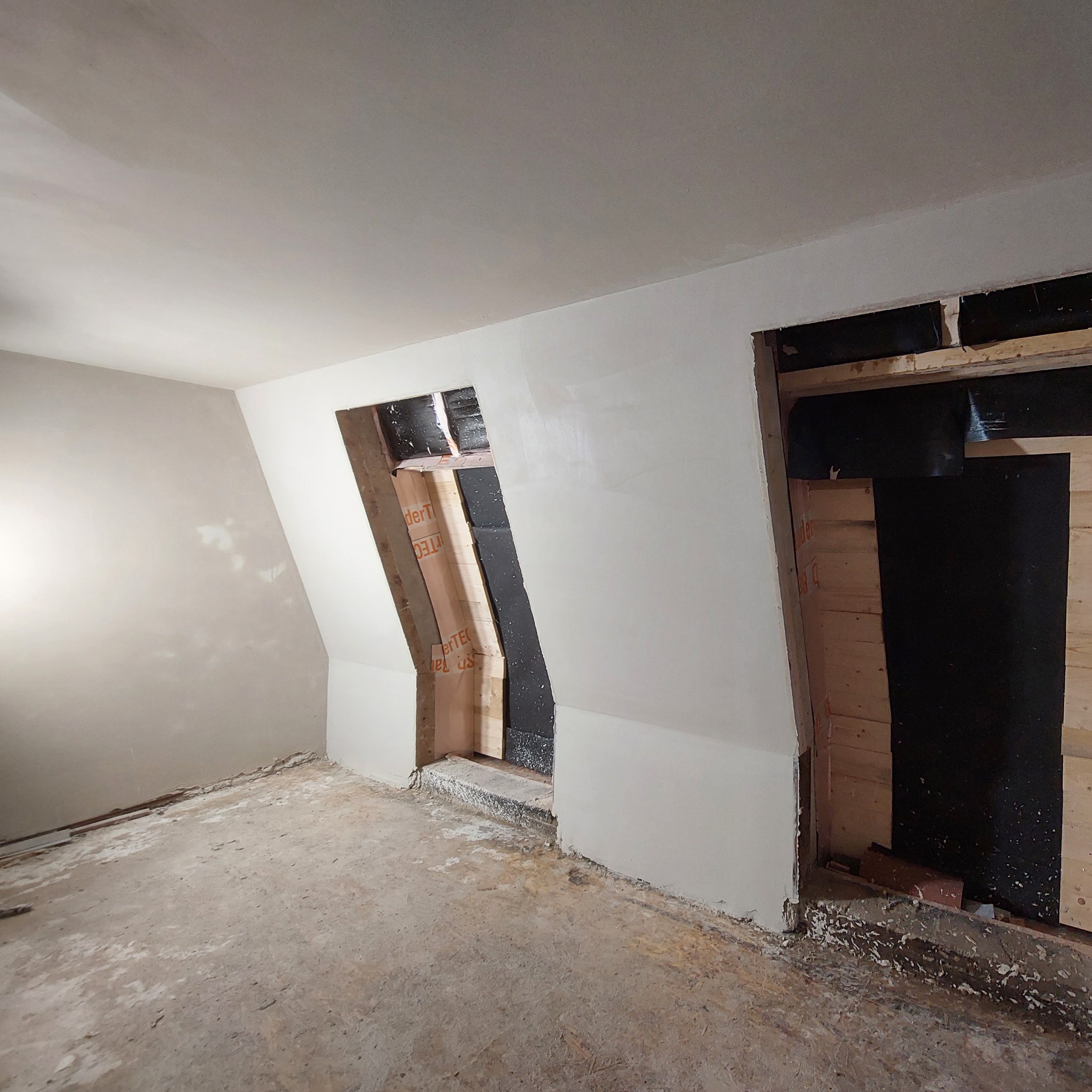 Wnętrze w trakcie prac wykończeniowych: ściany częściowo pokryte gładzią, widoczne ramy okienne z izolacją, surowy beton na podłodze.