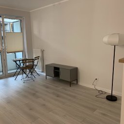 Happy Home - Jasne wnętrze po remoncie: panele podłogowe, szare meble, białe ściany i lampa. Balkon z meblami w tle. Widoczne gniazdko elektryczne.