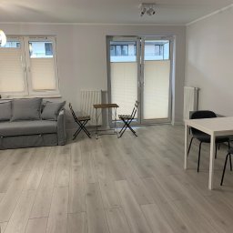 Happy Home - Jasne wnętrze pokoju z drewnianą podłogą, szarą sofą, stołem i krzesłami. Okno z żaluzjami plisowanymi. Minimalistyczny wystrój. Nowoczesna lampa wisząca.