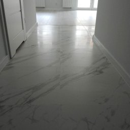 Happy Home - Błyszcząca, marmurowa podłoga z jasnymi płytkami w nowym mieszkaniu. Widoczne białe listwy przypodłogowe i fragmenty drzwi. W tle okno balkonowe i grzejnik.