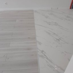 Happy Home - Połączenie paneli podłogowych w odcieniach szarości z płytkami imitującymi biały marmur na podłodze w jasnym wnętrzu. Widoczne listwy przypodłogowe.