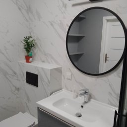 Happy Home - Nowoczesna łazienka z umywalką, okrągłym lustrem i marmurowymi płytkami. Minimalistyczny design z rośliną w doniczce jako akcent dekoracyjny.