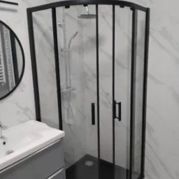 Nowoczesna kabina prysznicowa z czarnymi profilami na tle marmurowych płytek. Umywalka z szarą szafką. Okrągłe lustro w czarnej ramie dopełnia aranżację.