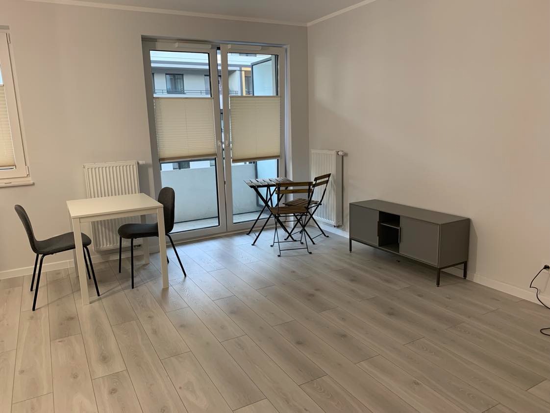 Jasne panele podłogowe w pokoju z białymi ścianami, stolikiem i krzesłami. Widok na balkon przez przeszklone drzwi z plisowanymi roletami.