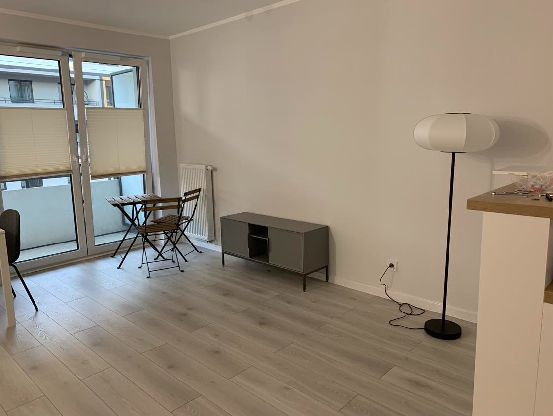 Jasne wnętrze po remoncie: panele podłogowe, szare meble, białe ściany i lampa. Balkon z meblami w tle. Widoczne gniazdko elektryczne.