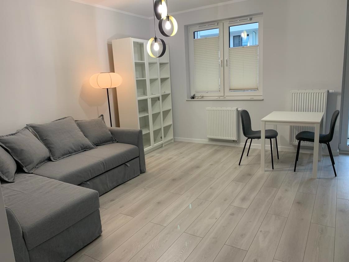 Jasne panele podłogowe w pokoju z szarą sofą narożną, białym regałem i stołem z krzesłami. Nowoczesne oświetlenie sufitowe i grzejniki ścienne.