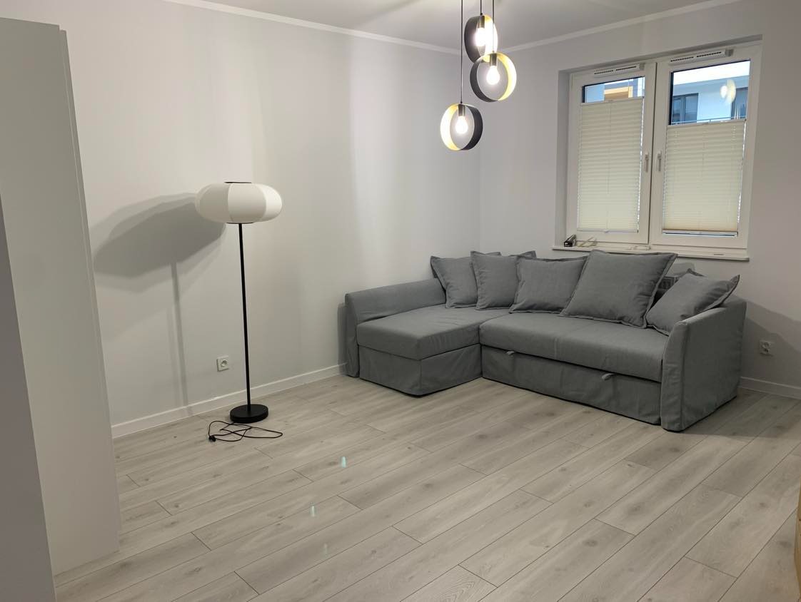 Jasne wnętrze pokoju z szarą sofą narożną, lampą podłogową i wiszącymi lampami. Widoczne panele podłogowe i okno z żaluzjami.