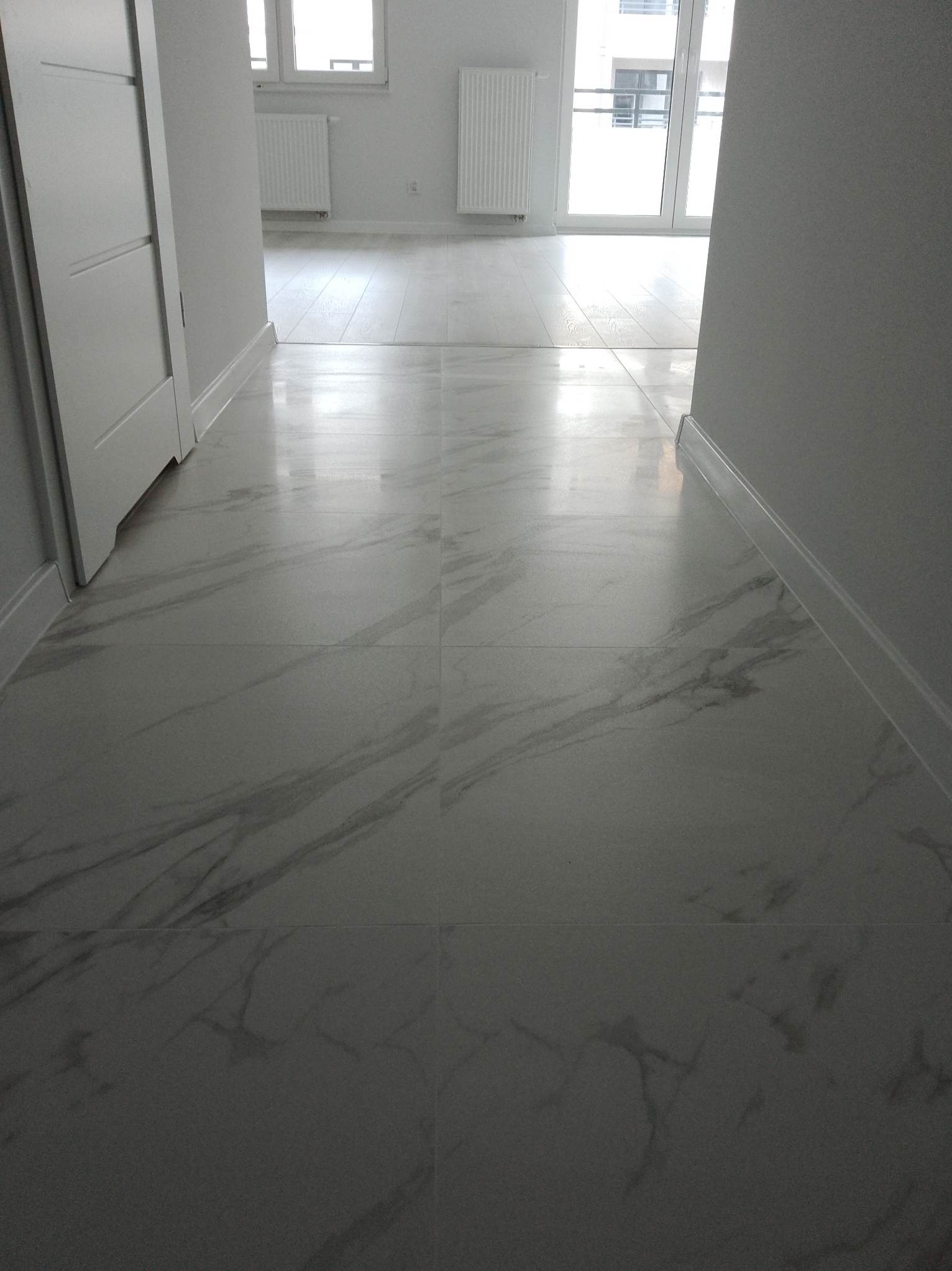 Błyszcząca, marmurowa podłoga z jasnymi płytkami w nowym mieszkaniu. Widoczne białe listwy przypodłogowe i fragmenty drzwi. W tle okno balkonowe i grzejnik.