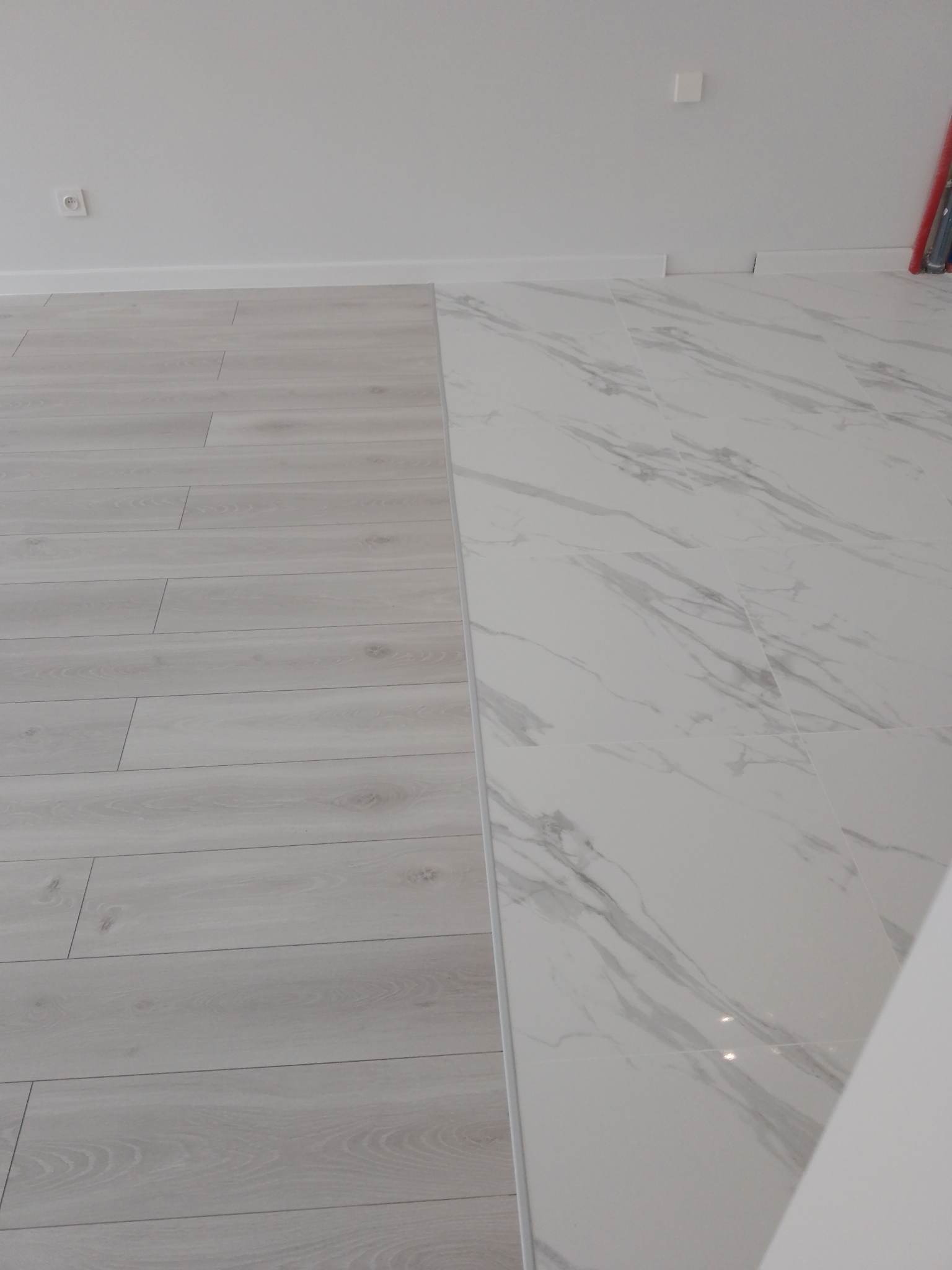 Połączenie paneli podłogowych w odcieniach szarości z płytkami imitującymi biały marmur na podłodze w jasnym wnętrzu. Widoczne listwy przypodłogowe.