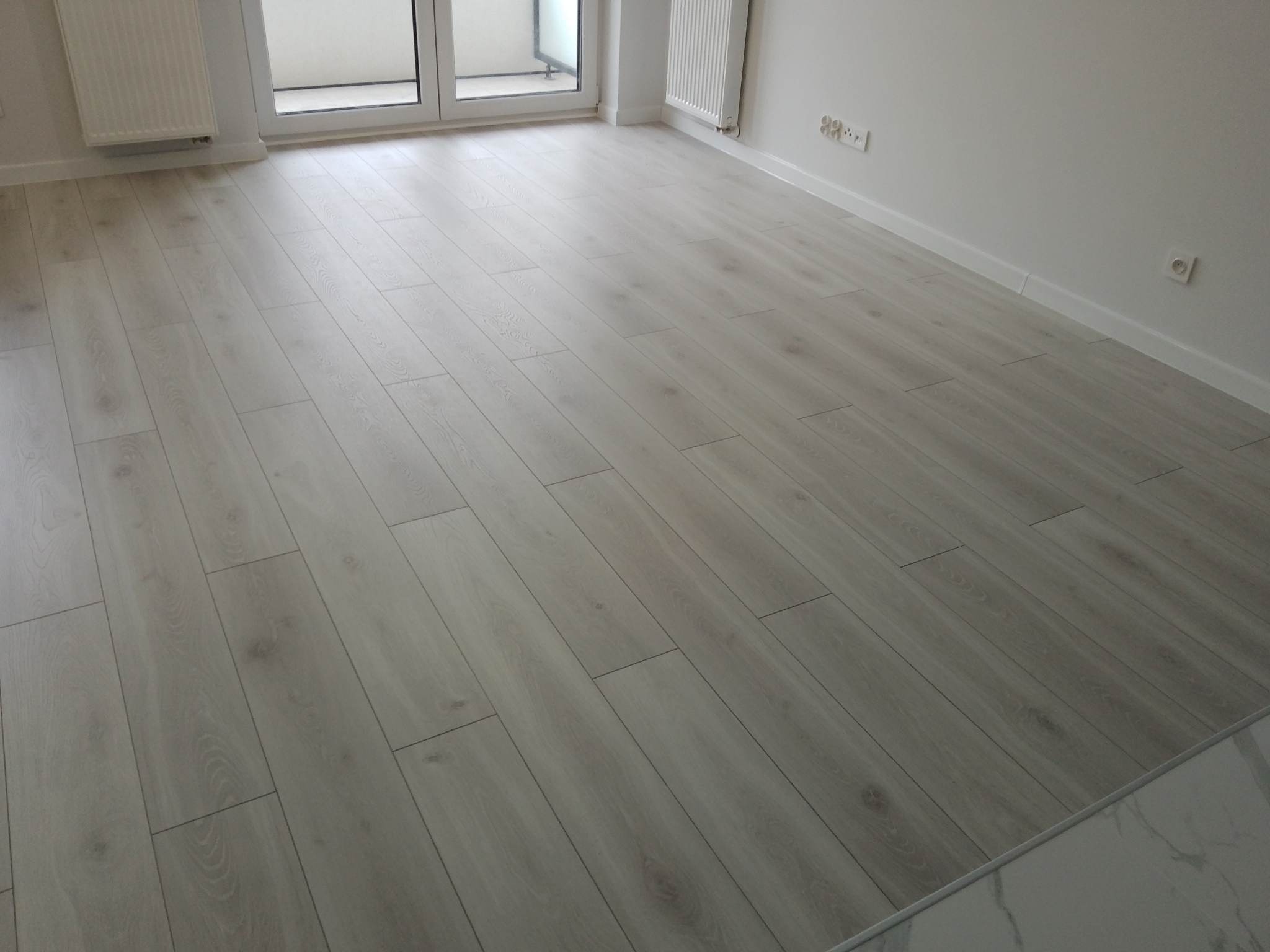 Jasne panele podłogowe ułożone w przestronnym wnętrzu z białymi ścianami, grzejnikiem i oknem balkonowym. Widoczne listwy przypodłogowe i gniazdka elektryczne.
