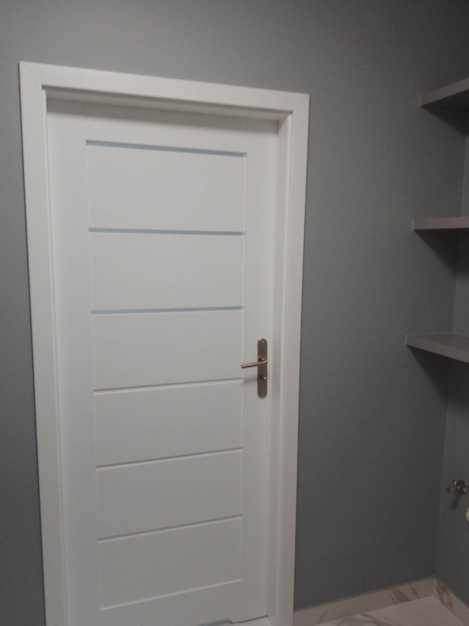 Białe, nowoczesne drzwi z poziomymi przeszkleniami w szarej łazience z półkami. Minimalistyczny design, jasne kolory, po remoncie.