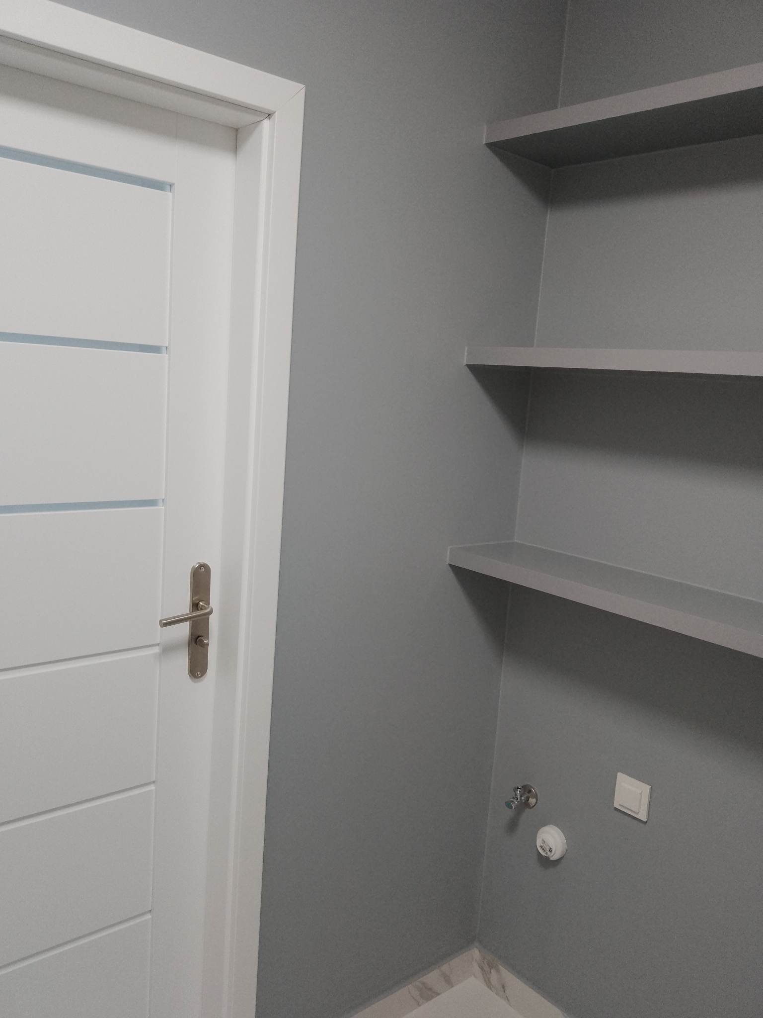Szara łazienka po remoncie: białe drzwi z poziomymi przeszkleniami, półki na ścianie, armatura i włącznik. Ściany w odcieniach szarości, marmurowe wykończenie podłogi.