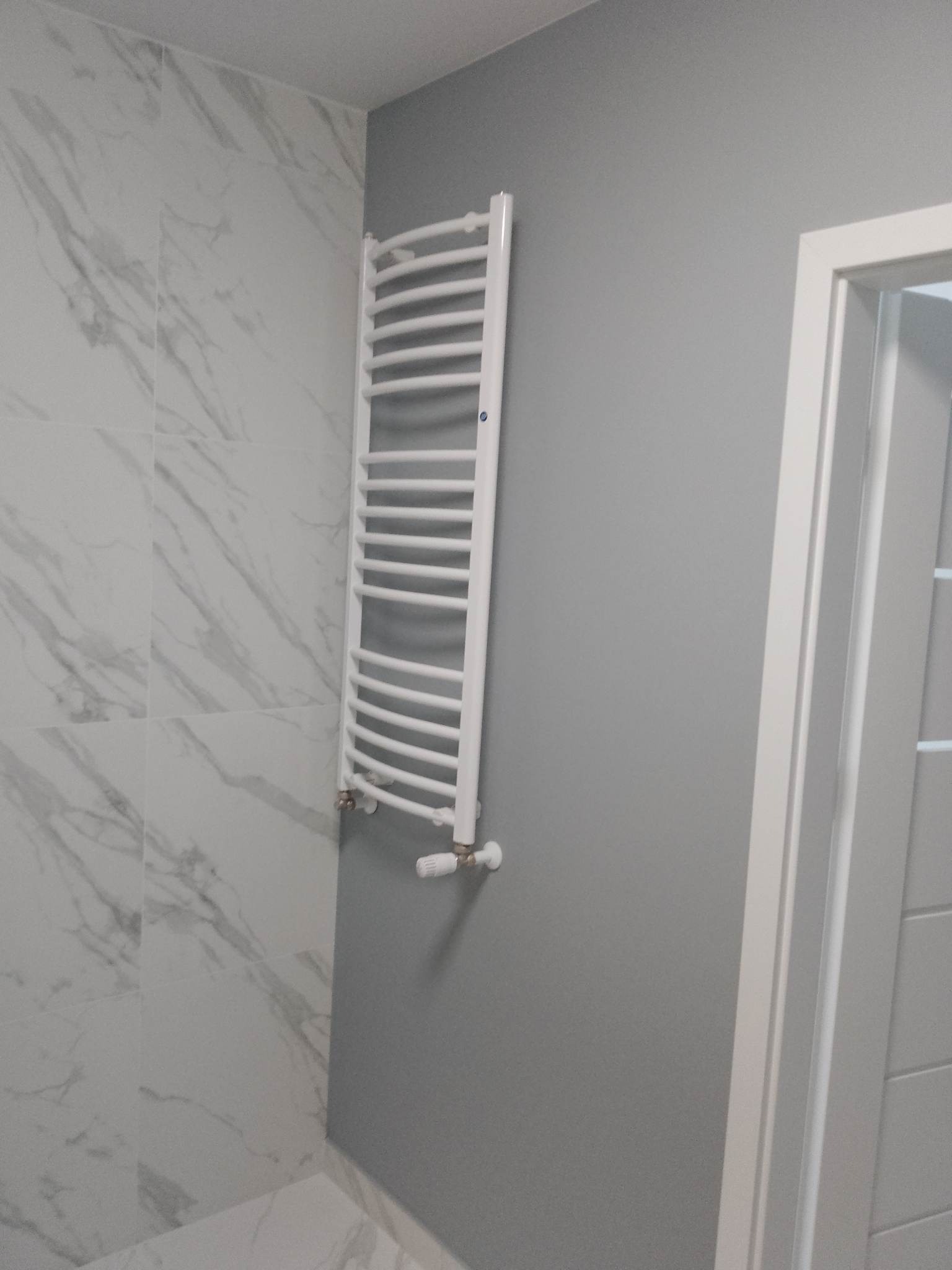 Biały grzejnik drabinkowy na szarej ścianie w łazience z marmurowymi płytkami. Widoczne detale wykończenia i fragment białych drzwi.