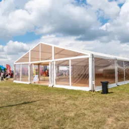 Długi, biały namiot z przezroczystymi ścianami na trawiastym terenie podczas targów, w tle widoczne traktory i inne stoiska.
