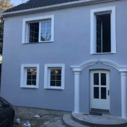 Dwukondygnacyjny dom z szarą elewacją, białymi obramowaniami okien i dekoracyjnym wejściem z kolumnami, widoczny samochód i akcesoria malarskie w pobliżu.