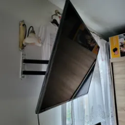 Telewizor LED zamontowany na ścianie na uchwycie, widoczny kabel zasilający, w tle lampa ścienna i firanka w oknie.