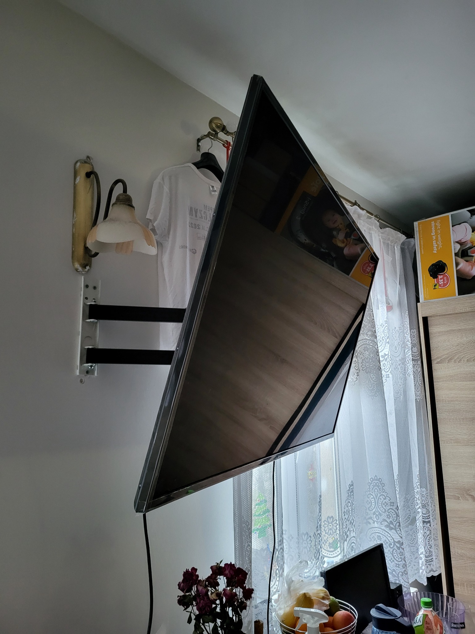 Telewizor LED zamontowany na ścianie na uchwycie, widoczny kabel zasilający, w tle lampa ścienna i firanka w oknie.