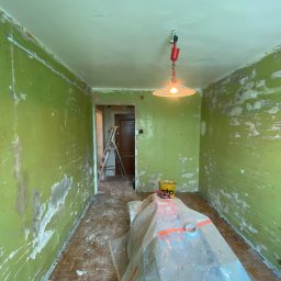 BudWolf Wykończenia Wnętrz Mateusz Wilk - Pokój w trakcie przygotowania do malowania: ściany z resztkami starej farby w odcieniach zieleni, miejscami zdarta, podłoga zabezpieczona folią malarską, widoczna drabina i wiadro z farbą.