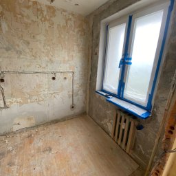 BudWolf Wykończenia Wnętrz Mateusz Wilk - Pokój w stanie surowym przed pracami wykończeniowymi: ściany ze śladami starej farby i tapet, zaklejone okno folią i taśmą malarską, odsłonięte przewody elektryczne, drewniana podłoga z resztkami...