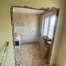 BudWolf Wykończenia Wnętrz Mateusz Wilk - Kuchnia przed remontem: zerwane tapety, odsłonięta cegła w miejscu planowanego przejścia, stara kuchenka i okno z widokiem na blok.