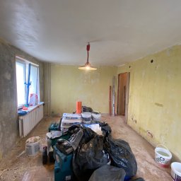 BudWolf Wykończenia Wnętrz Mateusz Wilk - Pokój przed remontem: widoczne stare, żółte ściany z ubytkami, okno z zabezpieczeniem folią, grzejnik, lampa wisząca, podłoga z resztkami materiałów budowlanych oraz worki z odpadami i narzędzia.