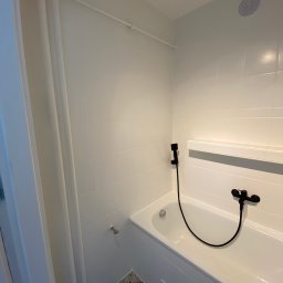 BudWolf Wykończenia Wnętrz Mateusz Wilk - Biała wanna z czarną armaturą i słuchawką prysznicową na tle białych płytek, z dekoracyjną podłogą w mozaikę.