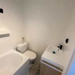 Nowoczesna, jasna łazienka po remoncie: białe płytki, umywalka z szafką z jasnego drewna, czarna bateria, toaleta i fragment wanny. Dekoracyjna podłoga z wzorzystych płytek.