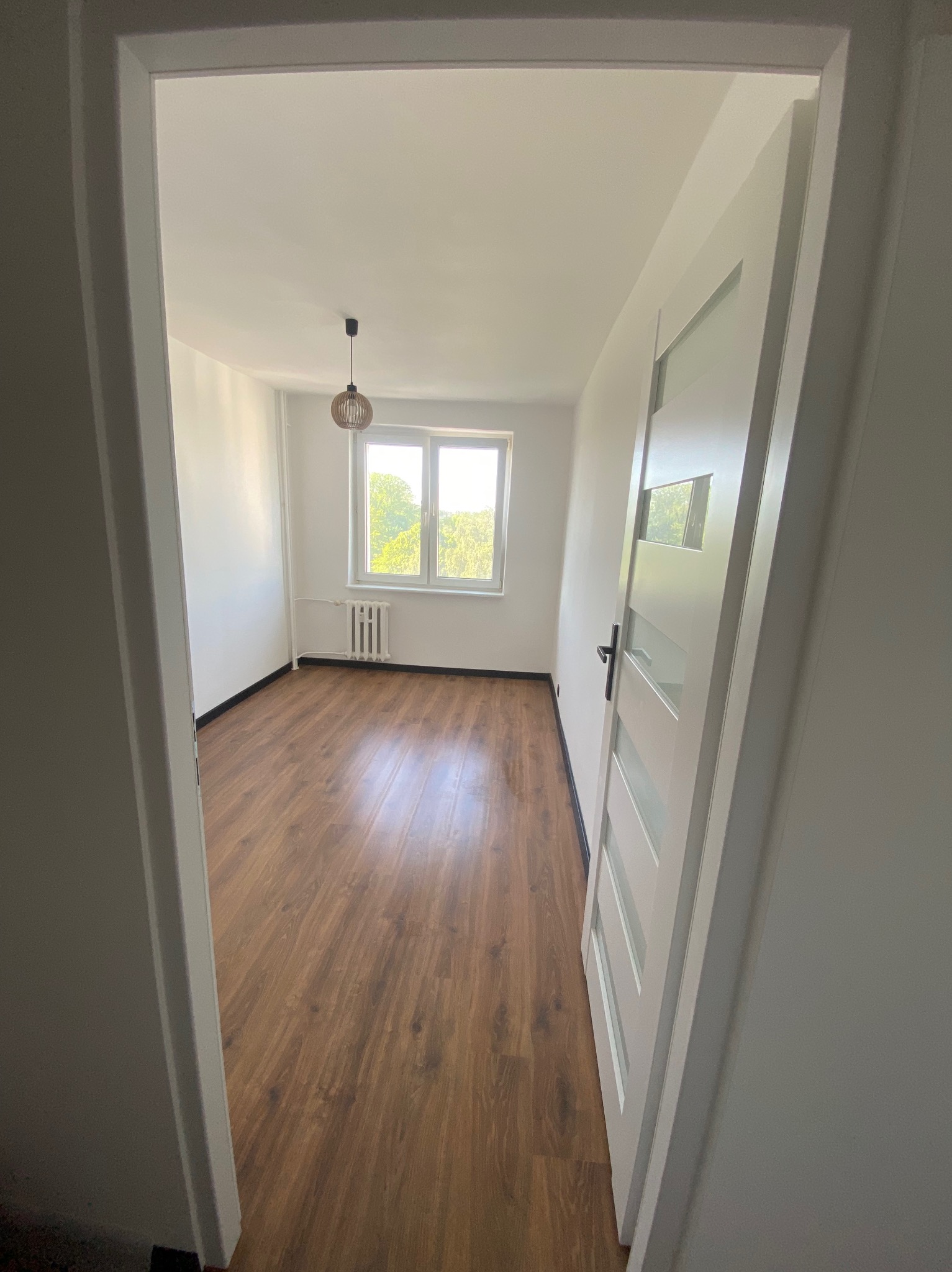 Widok zza otwartych drzwi na odnowione, puste pomieszczenie z drewnianą podłogą, białymi ścianami, grzejnikiem pod oknem i nowoczesną lampą wiszącą.