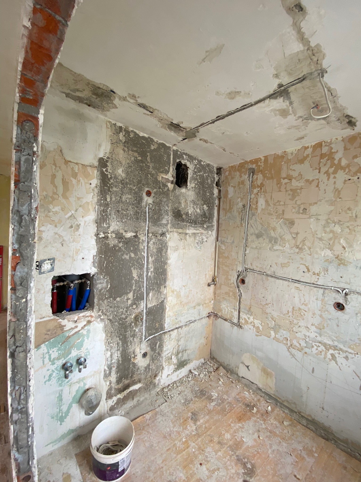 Łazienka w trakcie skuwania starych płytek i przygotowywania ścian pod remont, widoczne instalacje wodne i elektryczne, gruz na podłodze i wiadro z kablami.
