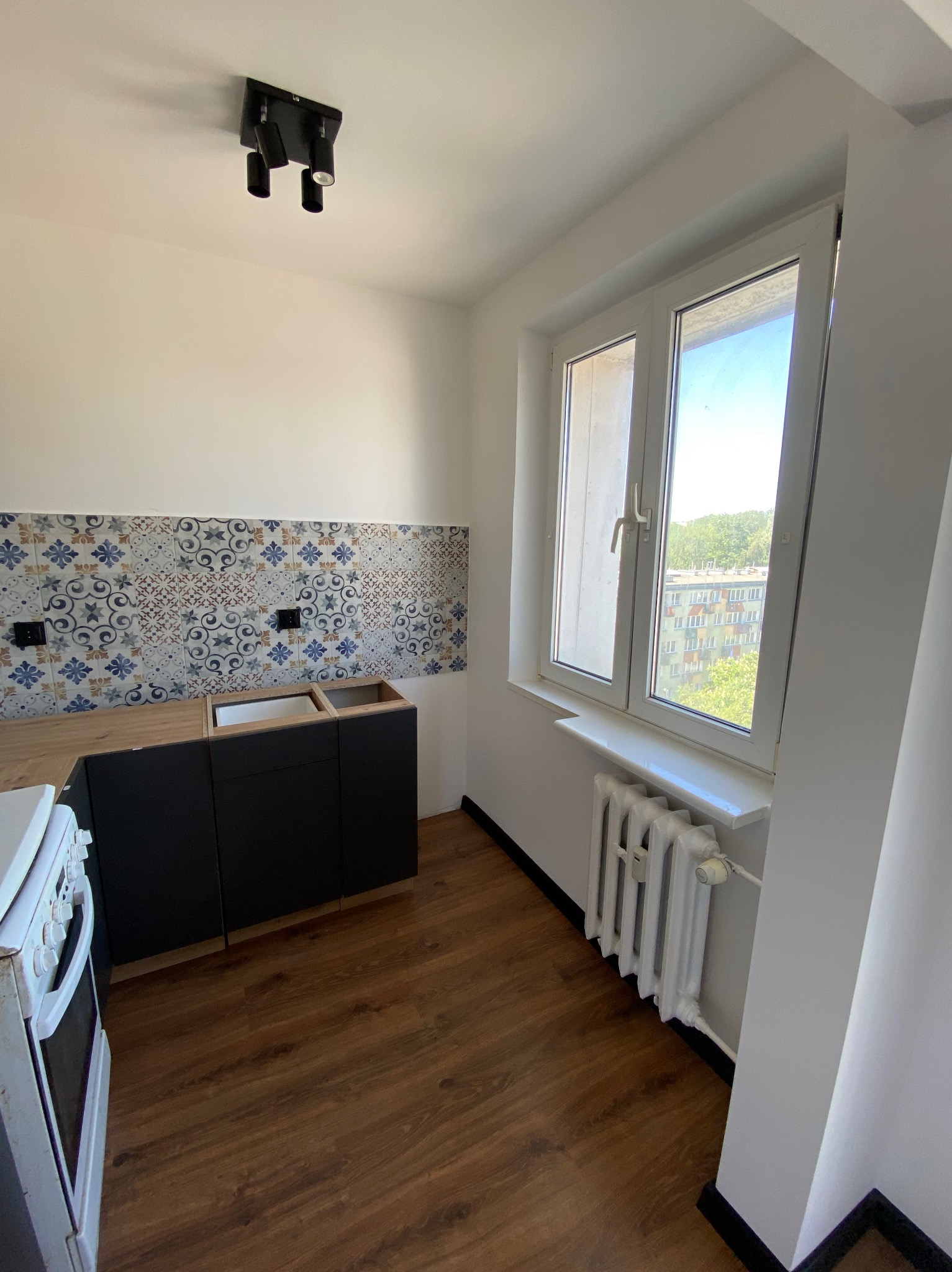 Kuchnia w trakcie montażu: czarne szafki bez frontów, blat z otworem na zlew, dekoracyjne płytki nad blatem, widok na blok przez okno.