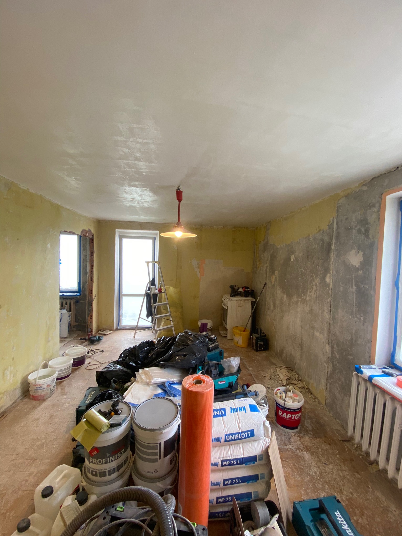 Pokój w trakcie prac przygotowawczych do remontu: częściowo otynkowane ściany, sufit po nałożeniu gładzi, widoczne worki z gładzią szpachlową Knauf, drabina aluminiowa i narzędzia malarskie.