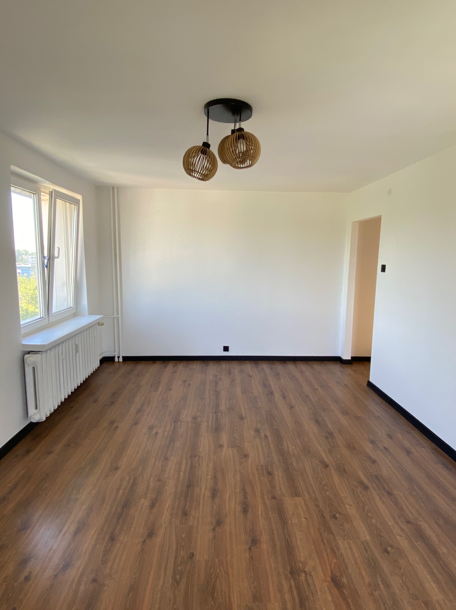 Pusty pokój po remoncie z drewnianą podłogą, białymi ścianami, grzejnikiem pod oknem i ozdobną lampą wiszącą.