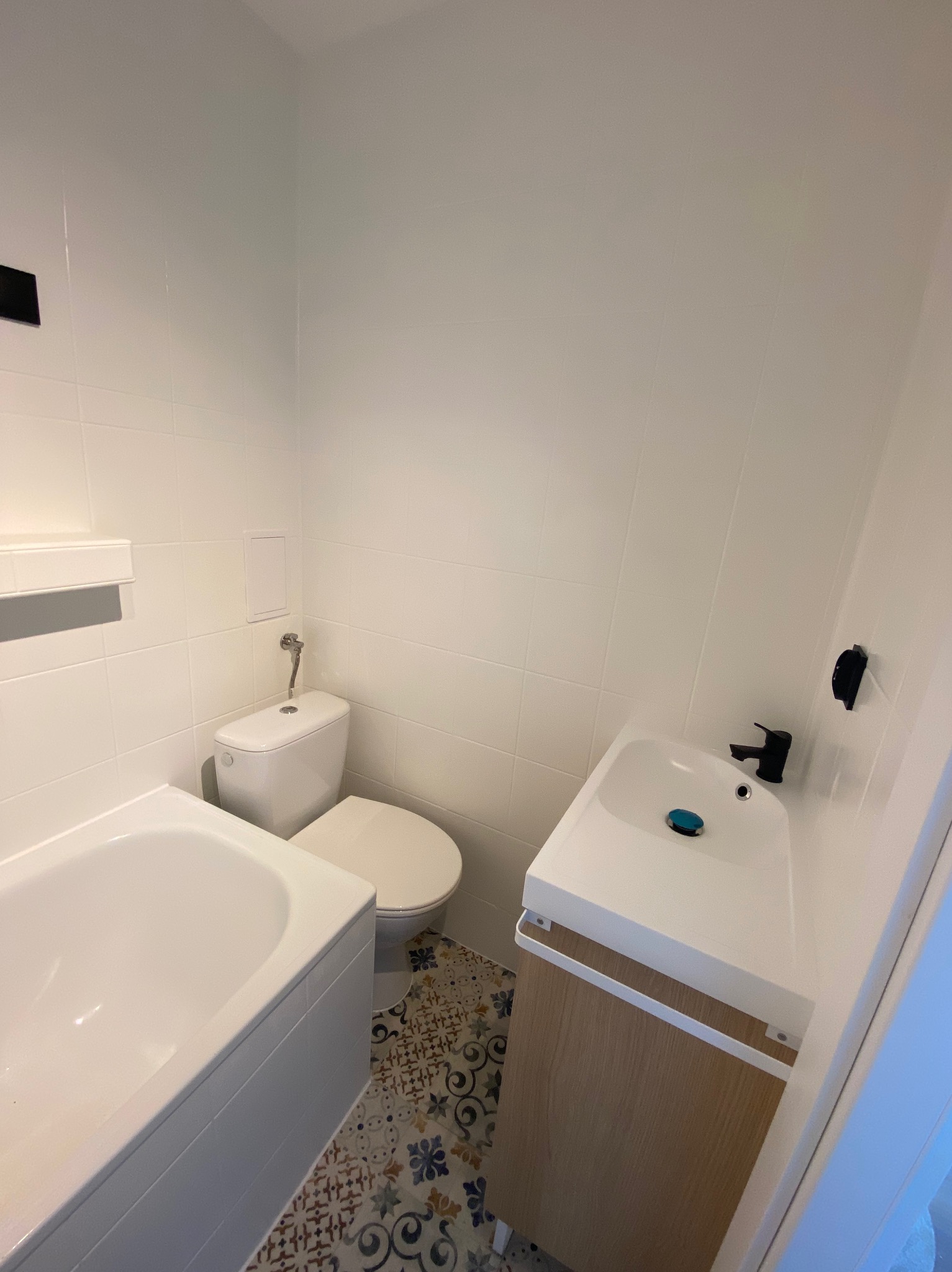 Nowoczesna, jasna łazienka po remoncie: białe płytki, umywalka z szafką z jasnego drewna, czarna bateria, toaleta i fragment wanny. Dekoracyjna podłoga z wzorzystych płytek.