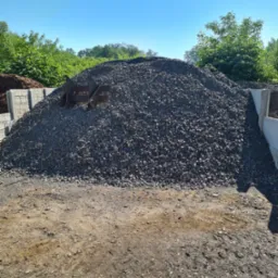 Kupa drobnego, szarego kamienia na placu budowy, widoczne zęby łyżki koparki i betonowe elementy ograniczające składowisko.