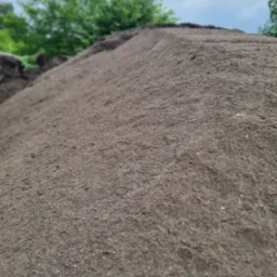 Duża pryzma ciemnej, wilgotnej ziemi z widocznymi grudkami i drobnymi kamykami, w tle zieleń drzew i pochmurne niebo.