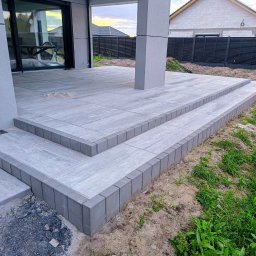 Ogród i Kamień - Nowoczesny taras z szarych płyt betonowych i obrzeży, z dwoma stopniami, przylegający do budynku z dużymi oknami przesuwnymi. Widoczne trawnik i ogrodzenie.