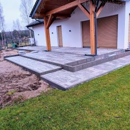 Ogród i Kamień - Ułożona kostka brukowa w odcieniach szarości tworząca taras z schodami przy domu z drewnianą pergolą. Widoczne trawnik i przygotowany teren pod dalsze prace.