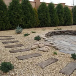 Ogród z ścieżką z drewnianych płyt ułożonych na żwirze, z dekoracyjnym murkiem z kamienia łupanego i żywopłotem z tuj w tle.