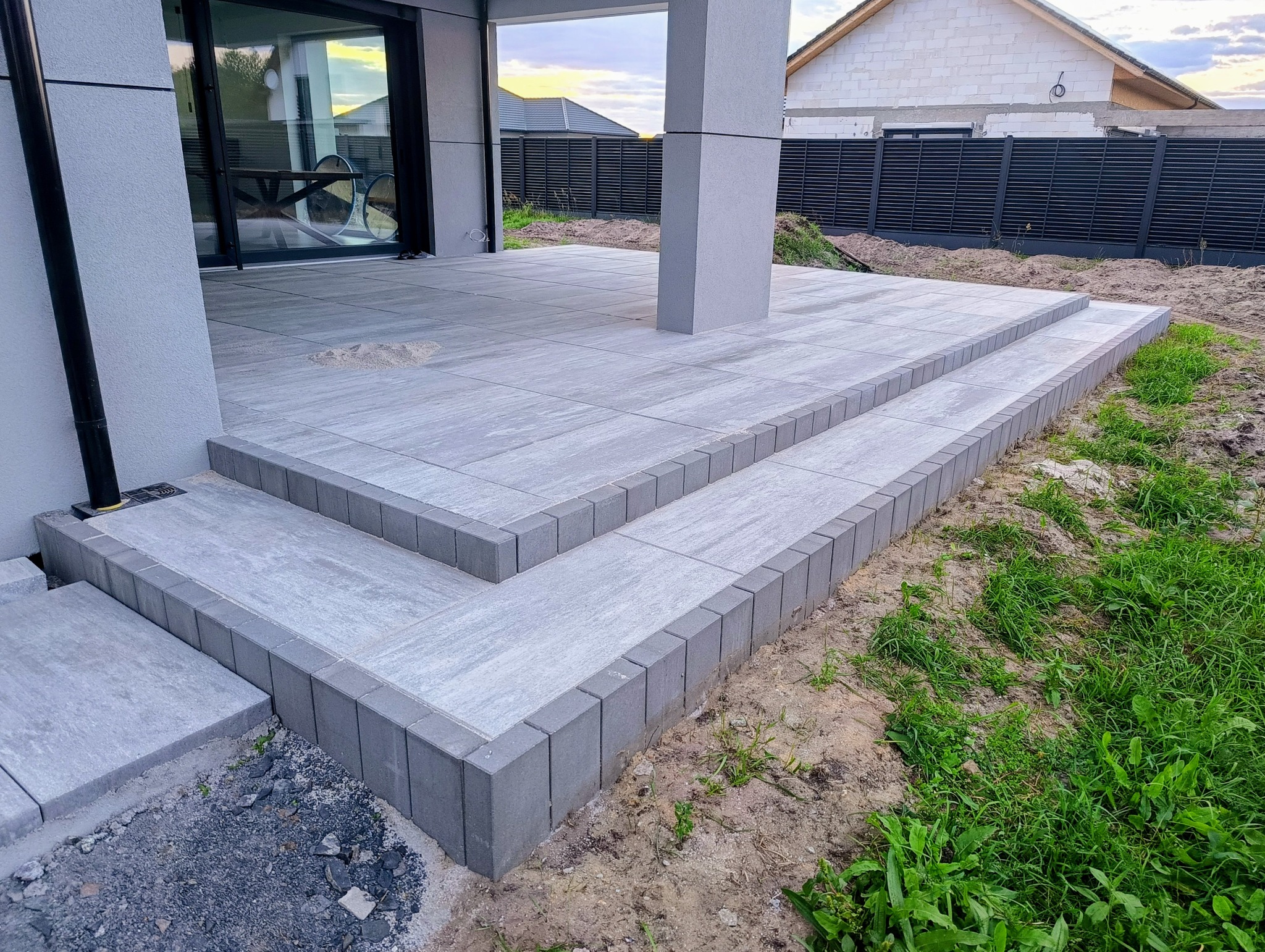 Nowoczesny taras z szarych płyt betonowych i obrzeży, z dwoma stopniami, przylegający do budynku z dużymi oknami przesuwnymi. Widoczne trawnik i ogrodzenie.