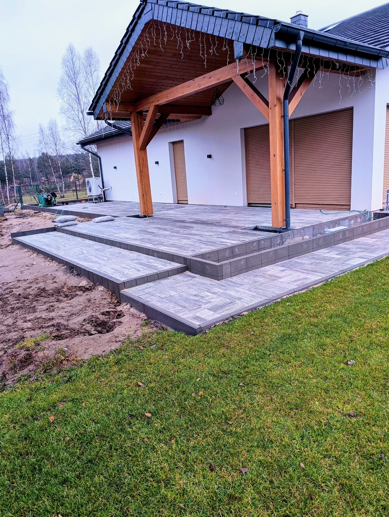 Ułożona kostka brukowa w odcieniach szarości tworząca taras z schodami przy domu z drewnianą pergolą. Widoczne trawnik i przygotowany teren pod dalsze prace.