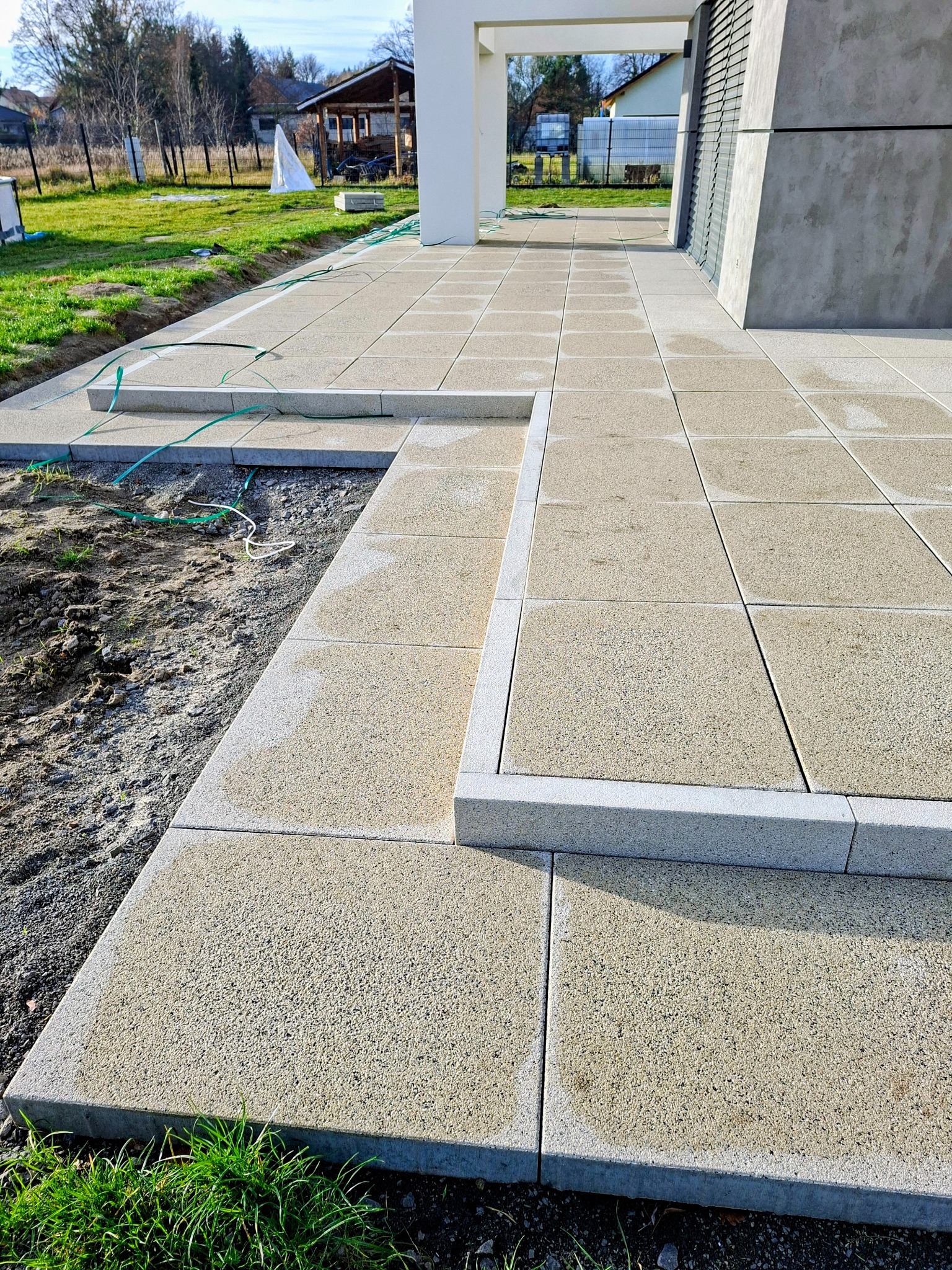 Ułożona kostka brukowa w odcieniach szarości tworząca ścieżkę z niskimi schodami. Widoczne elementy domu i trawnik w tle. Świeżo wykonana nawierzchnia.