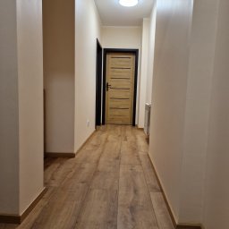 Marek Kochajkiewicz - Korytarz po remoncie z drewnopodobną podłogą, świeżo pomalowanymi ścianami w odcieniach beżu i nowymi drzwiami z czarną ościeżnicą oraz grzejnikiem.