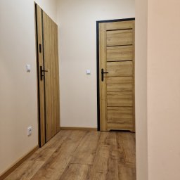 Marek Kochajkiewicz - Korytarz z dwiema drewnianymi drzwiami z czarnymi obramowaniami i klamkami, parkiet na podłodze, jasne ściany, widoczna instalacja elektryczna.