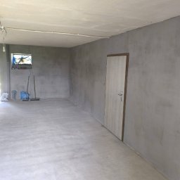 Marek Kochajkiewicz - Szara, pusta przestrzeń po gładziowaniu ścian i wylewce podłogi, z widocznymi narzędziami i zamontowanymi drzwiami. Okno z lewej strony doświetla wnętrze.