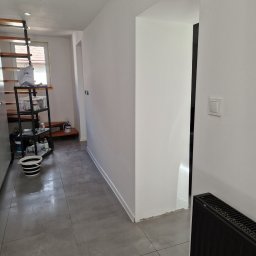 Marek Kochajkiewicz - Świeżo pomalowane, białe ściany w korytarzu z szarą podłogą, widoczny grzejnik i fragment regału z akcesoriami malarskimi. Oświetlenie sufitowe.