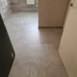 Wykończona łazienka z szarymi płytkami na podłodze i ścianach. Widoczny biały grzejnik na ścianie i fragment otwartych drzwi po prawej stronie. Minimalistyczny design.