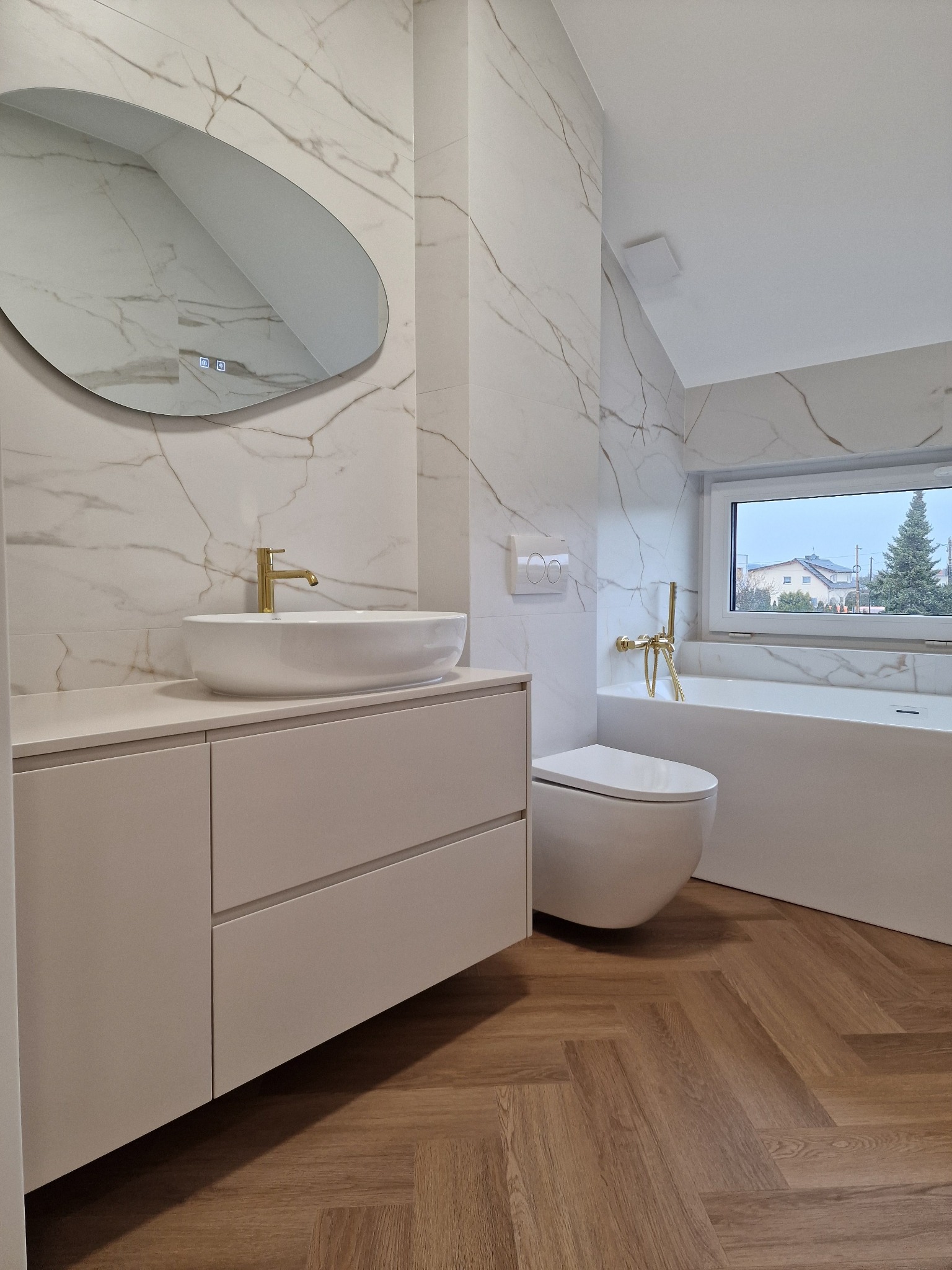 Nowoczesna łazienka z białymi marmurkowymi płytkami, umywalką nablatową na szafce z szufladami, podwieszaną toaletą i wanną przy oknie z widokiem na domy i drzewa. Złota armatura dodaje elegancji.
