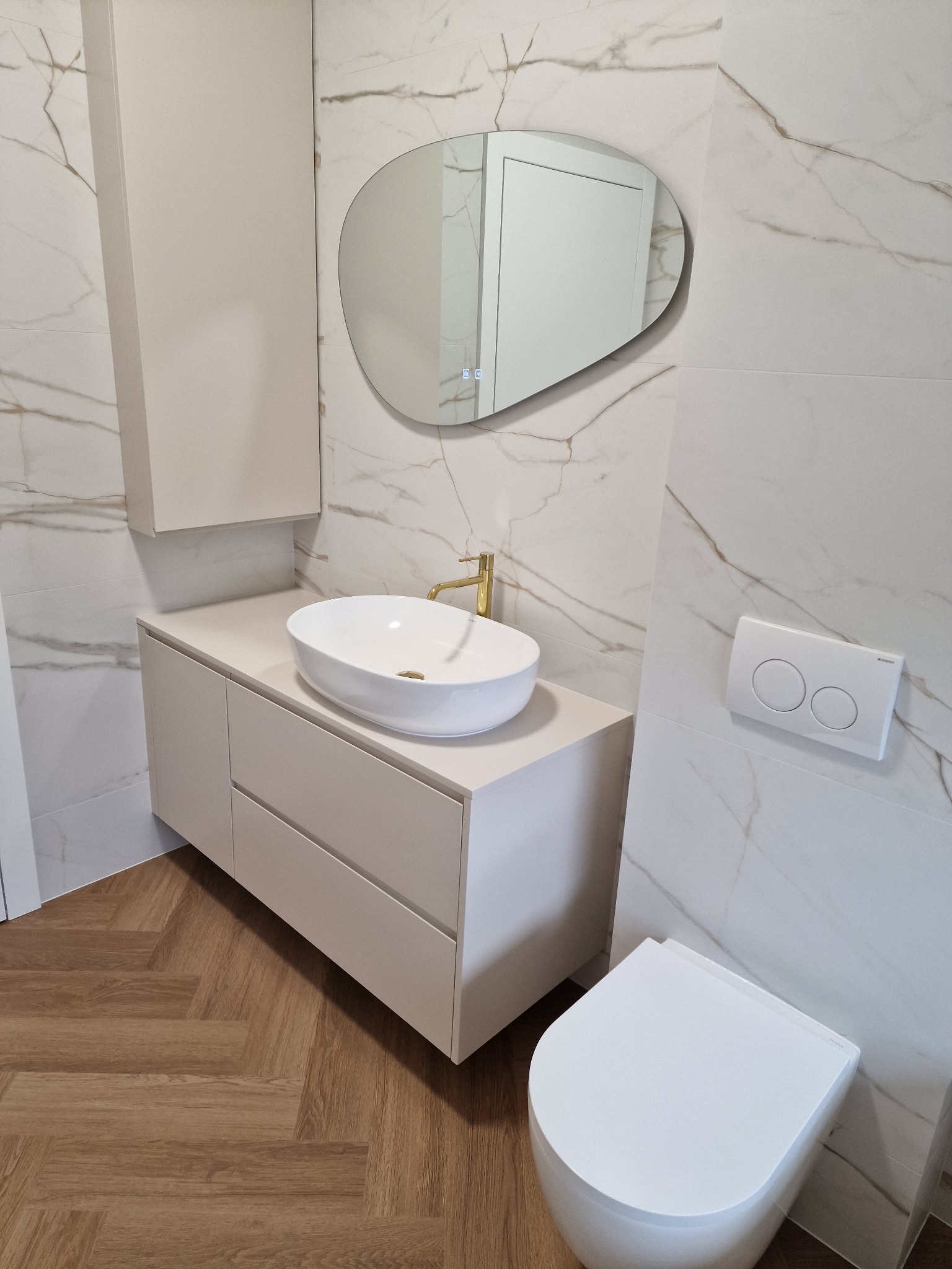 Nowoczesna łazienka z białymi marmurowymi płytkami, owalną umywalką na szafce z szufladami, asymetrycznym lustrem z podświetleniem LED i toaletą podwieszaną.