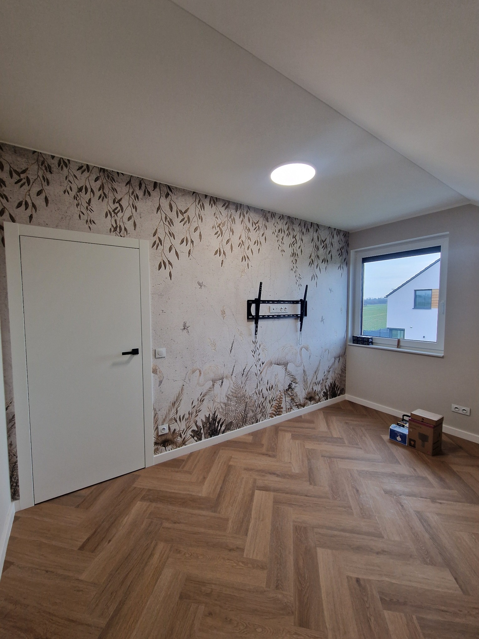 Pokój po remoncie z drewnianą podłogą ułożoną w jodełkę, ściana z tapetą w motywy roślinne i zwierzęce, uchwyt na telewizor na ścianie, białe drzwi i okno z widokiem na dom.