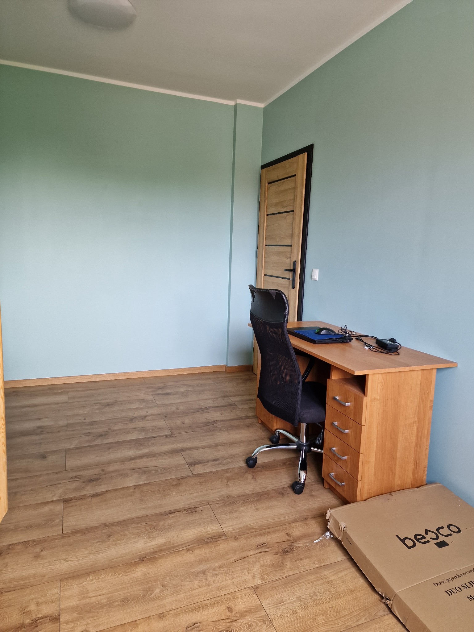 Pomalowane na błękitno ściany pokoju z drewnianą podłogą, biurkiem z krzesłem i zamkniętymi drzwiami. Na podłodze leży karton po drzwiach.